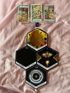 Three tarot cards - Two of Wands Rx, Seven of Pentacles, and King of Pentacles are in a straight line above a cross shaped layout of four Alchemy cards - The Despicable Face, The Void, Mystical Sister, and Synchronicty
